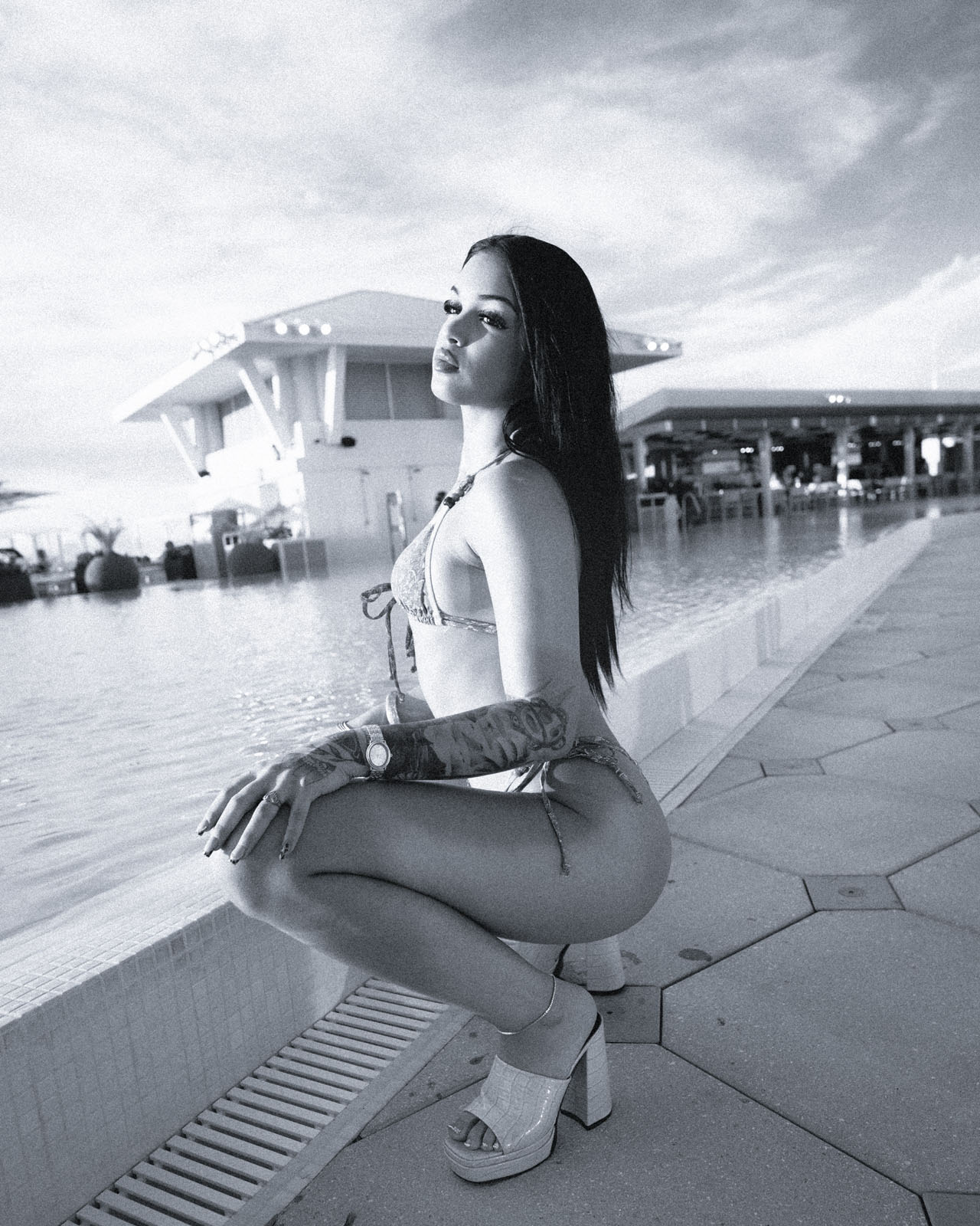 Black-and-white portrait of a tattooed woman crouching by a resort pool